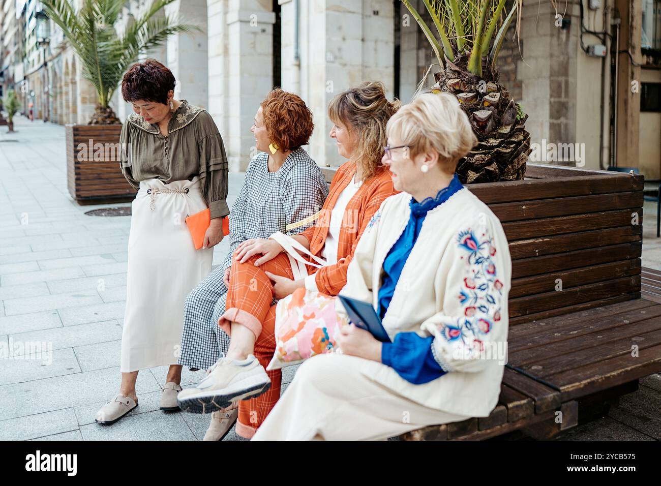 Quatre femmes de styles et d'apparences variés s'engagent dans une discussion animée tandis que trois d'entre elles sont assises sur un banc de rue, montrant une scène de frien Banque D'Images