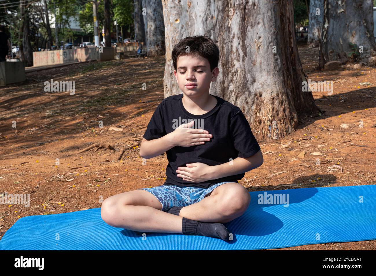 Enfant brésilien assis sur un tapis de yoga avec les mains sur la poitrine et l'abdomen en classe extérieure 4. Banque D'Images