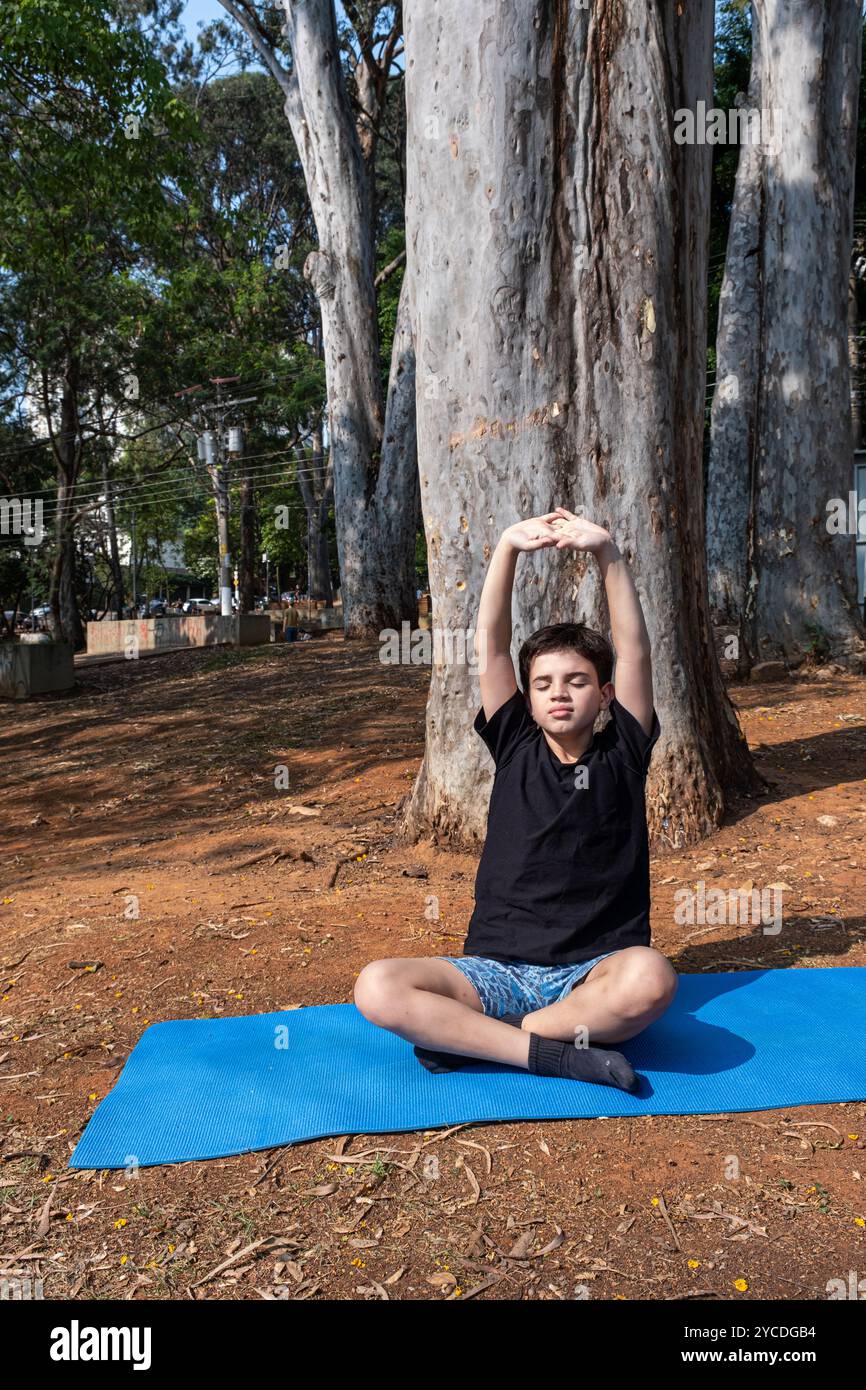 Enfant brésilien assis sur un tapis de yoga et étirant en classe extérieure 7. Banque D'Images