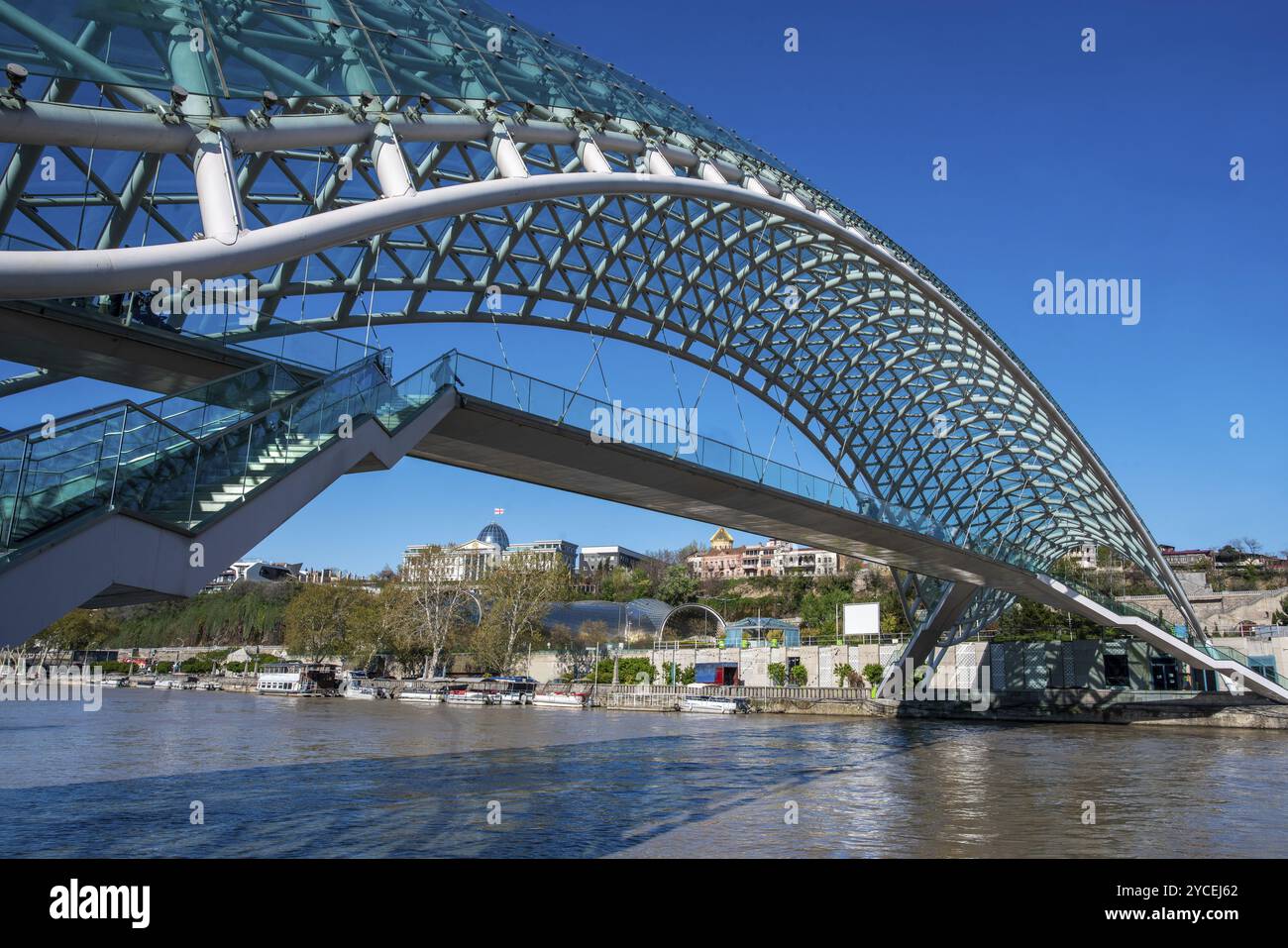 Pont piétonnier de la paix traversant la rivière Kura à Tbilissi. Au loin, le Palais présidentiel avec le drapeau géorgien dans un beau ciel bleu. Le Banque D'Images