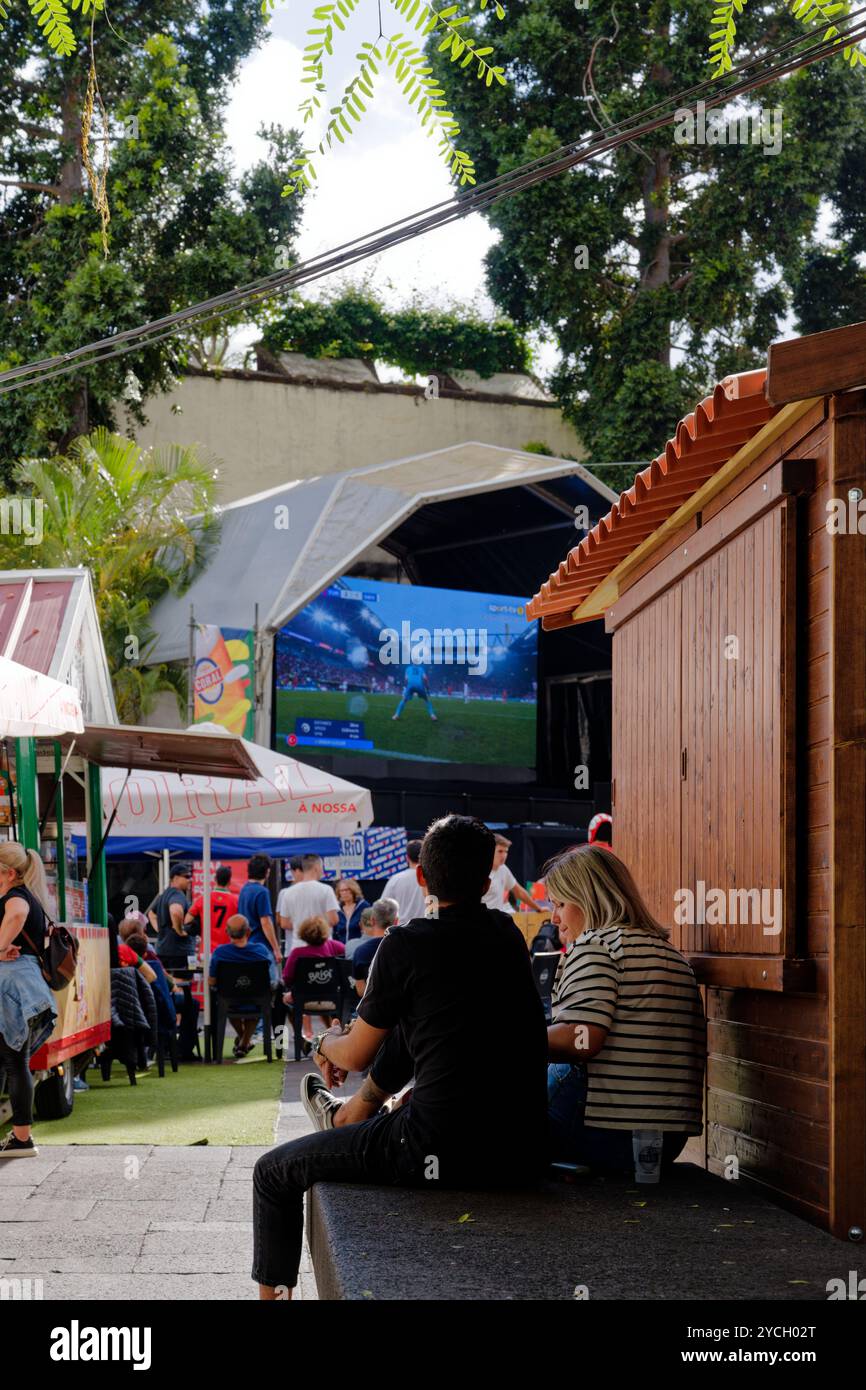 Couple à Funchal regardant un match de football en direct sur un grand écran extérieur, atmosphère détendue au milieu des stands de nourriture et des arbres ombragés Banque D'Images