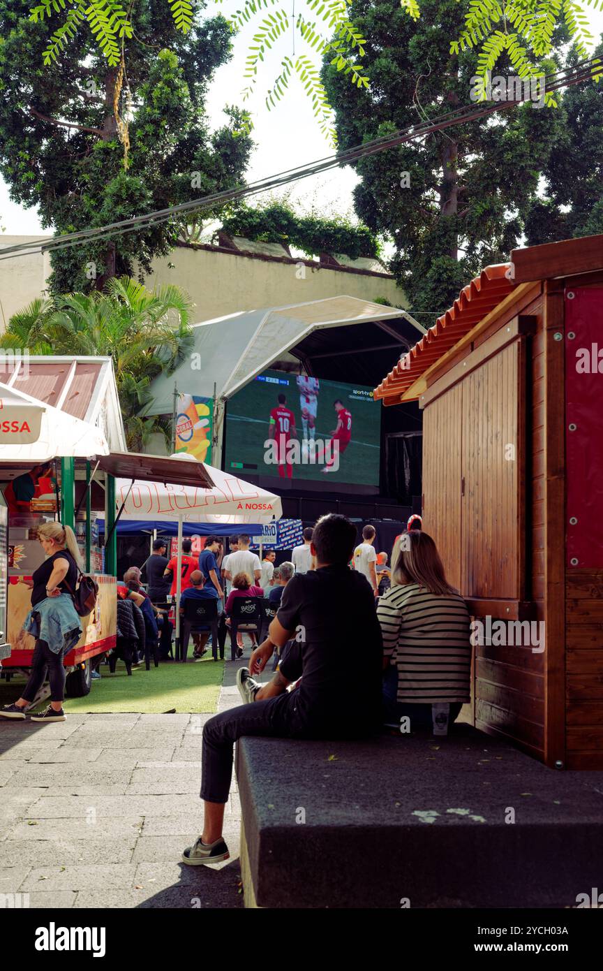 Couple à Funchal regardant un match de football en direct sur un grand écran extérieur, atmosphère détendue au milieu des stands de nourriture et des arbres ombragés Banque D'Images