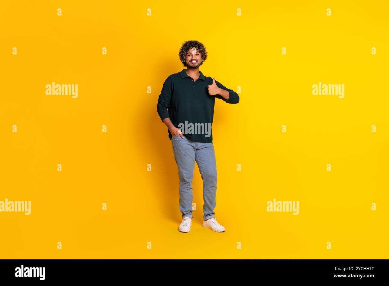 Un jeune homme portant une chemise noire et un Jean donne un pouce vers le haut sur un fond jaune vif, mettant en valeur une attitude joyeuse et positive Banque D'Images