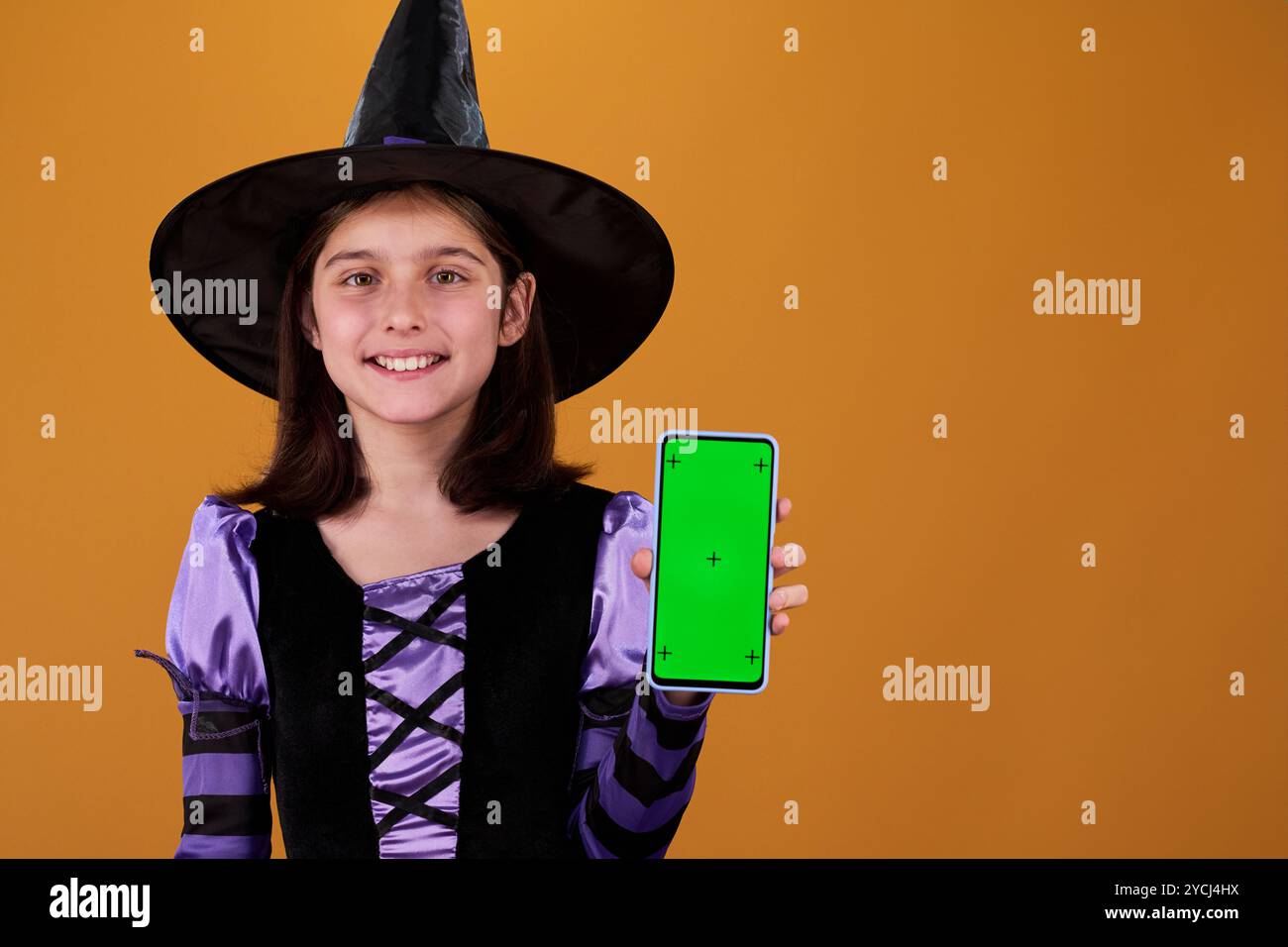 Fille mignonne heureuse en costume de sorcière d'Halloween vous montrant smartphone avec écran chromakey avec annonce de vente Banque D'Images
