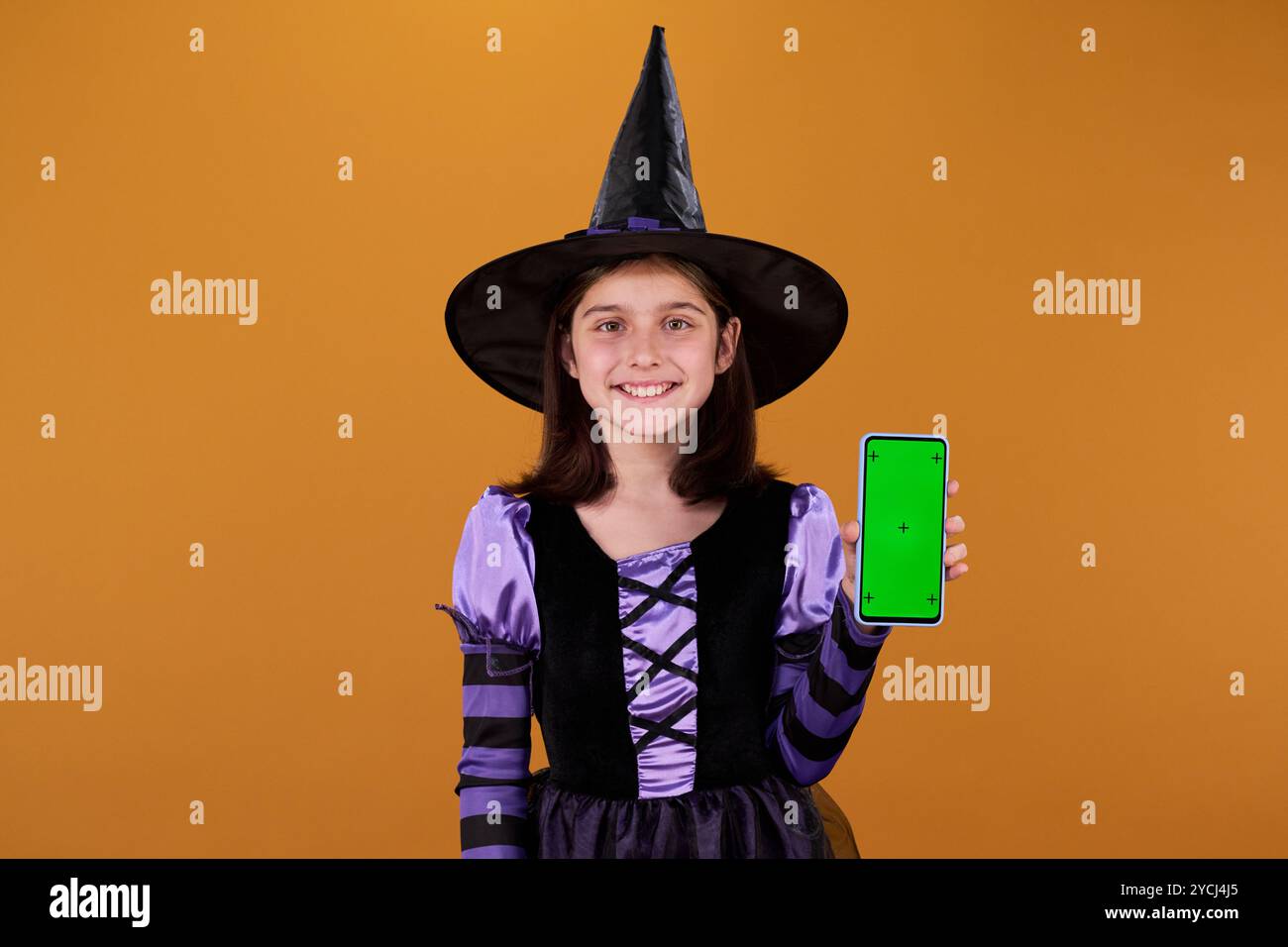 Fille souriante dans le costume d'Halloween de sorcière vous regardant et montrant l'écran vert de gadget mobile avec votre annonce Banque D'Images