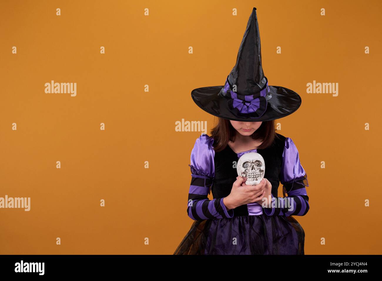 Sorcière jeune dans un grand chapeau noir et robe de fantaisie regardant le crâne humain dans ses mains tout en se tenant debout sur fond orange Banque D'Images