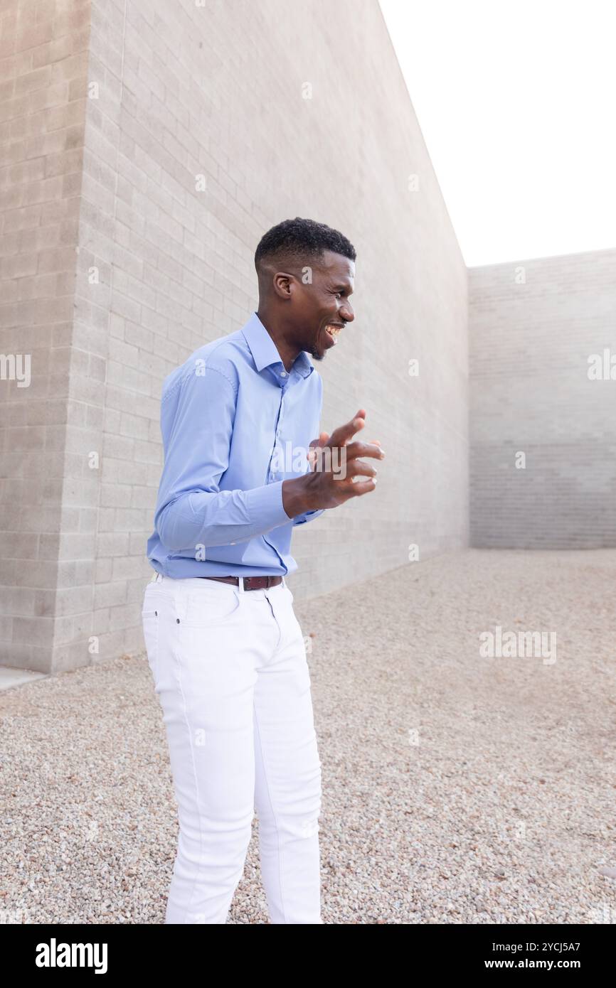 Rire homme afro-américain élégant portant un Jean blanc et une chemise bleue en plein air, mur de béton sur fond. Homme moyen âge à la mode dans ses années 40 Banque D'Images