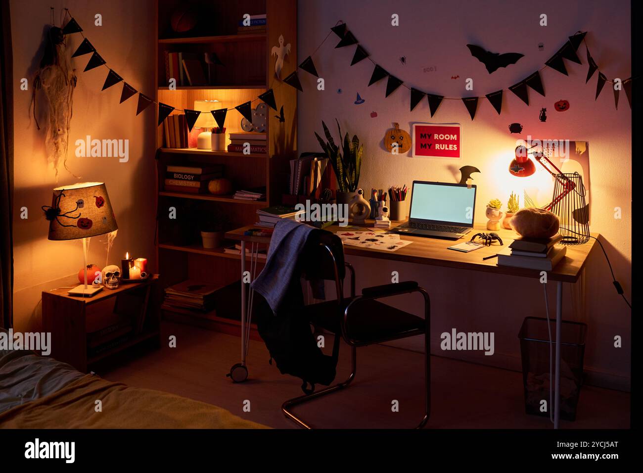 Une partie de la chambre domestique spacieuse avec chaise debout devant la table avec ordinateur portable, symboles d'Halloween, livres, lampe et autres choses Banque D'Images