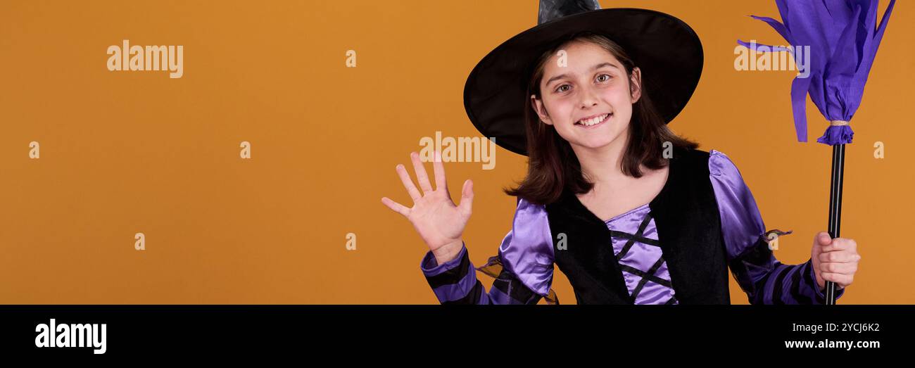En-tête de fille mignonne dans le costume d'Halloween de sorcière tenant le balai et agitant la main à vous tout en restant debout par copyspace pour votre annonce Banque D'Images