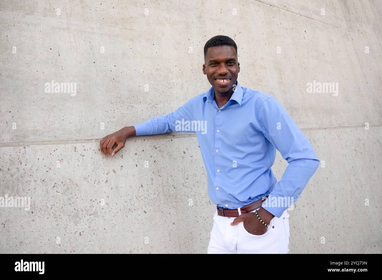 Portrait de beau sourire avec des dents homme afro-américain portant Jean blanc et chemise bleue, ceinture brune extérieure, mur de béton sur fond. Copier Banque D'Images