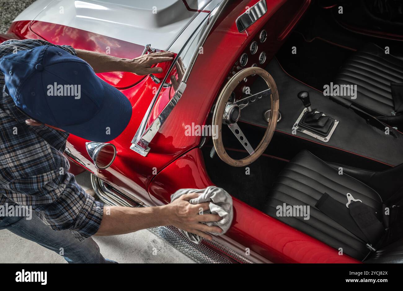 Une personne polit la surface brillante d'une voiture de sport rouge vintage dans un garage. La lumière du soleil met en valeur les détails de la voiture, créant une atmosphère chaleureuse. Banque D'Images