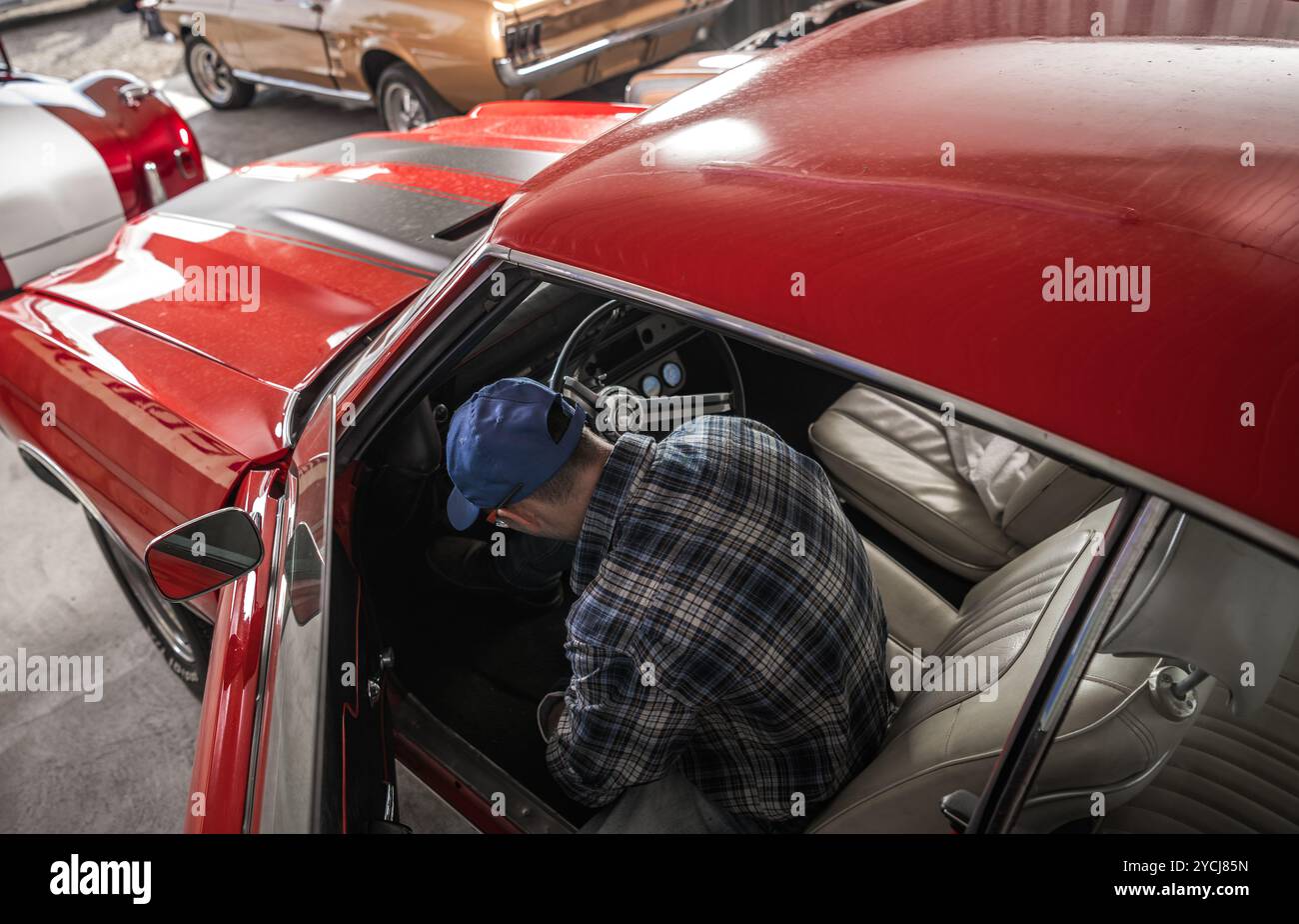 Un homme inspecte l'intérieur d'une voiture rouge classique, en se concentrant sur le tableau de bord tout en étant entouré d'autres voitures anciennes dans un garage bien éclairé Banque D'Images