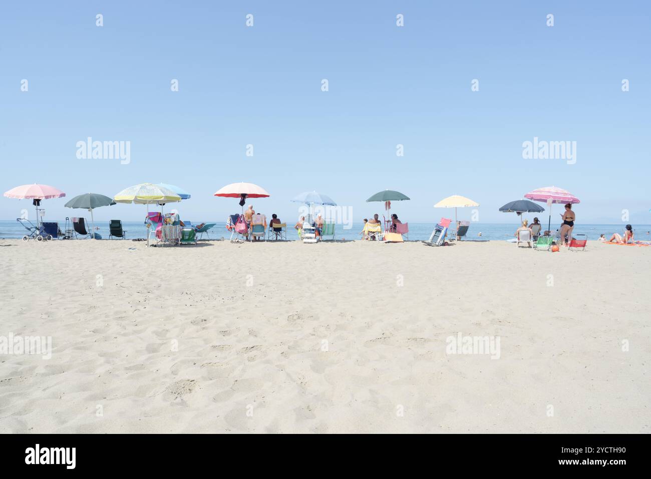 Un groupe de personnes debout en ligne sur une rive sablonneuse, face à l'eau calme, avec un ciel matinal délicat peint dans des tons pastel doux de rose et bleu Banque D'Images