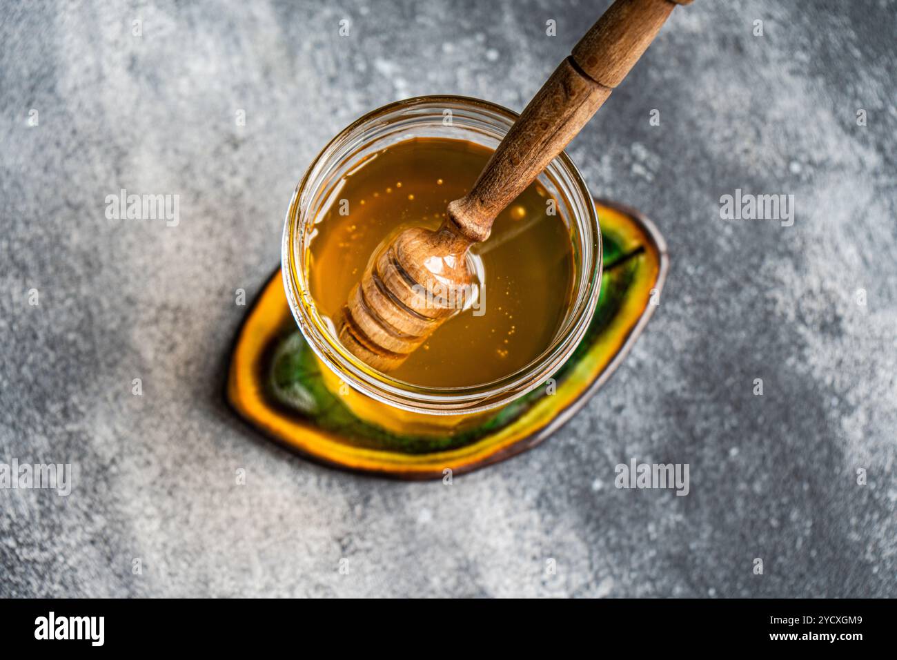 Vue de dessus de miel biologique doré dans un bocal en verre transparent, avec une trempette à miel en bois partiellement immergée. Capturé sur une surface grise texturée avec un Banque D'Images