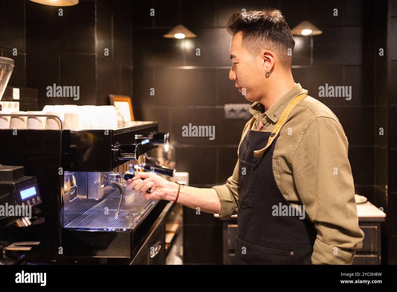 Asian Barista in Apron exploite habilement une machine à café dans un café moderne au thème sombre, mettant en valeur son expertise dans la préparation du café Banque D'Images