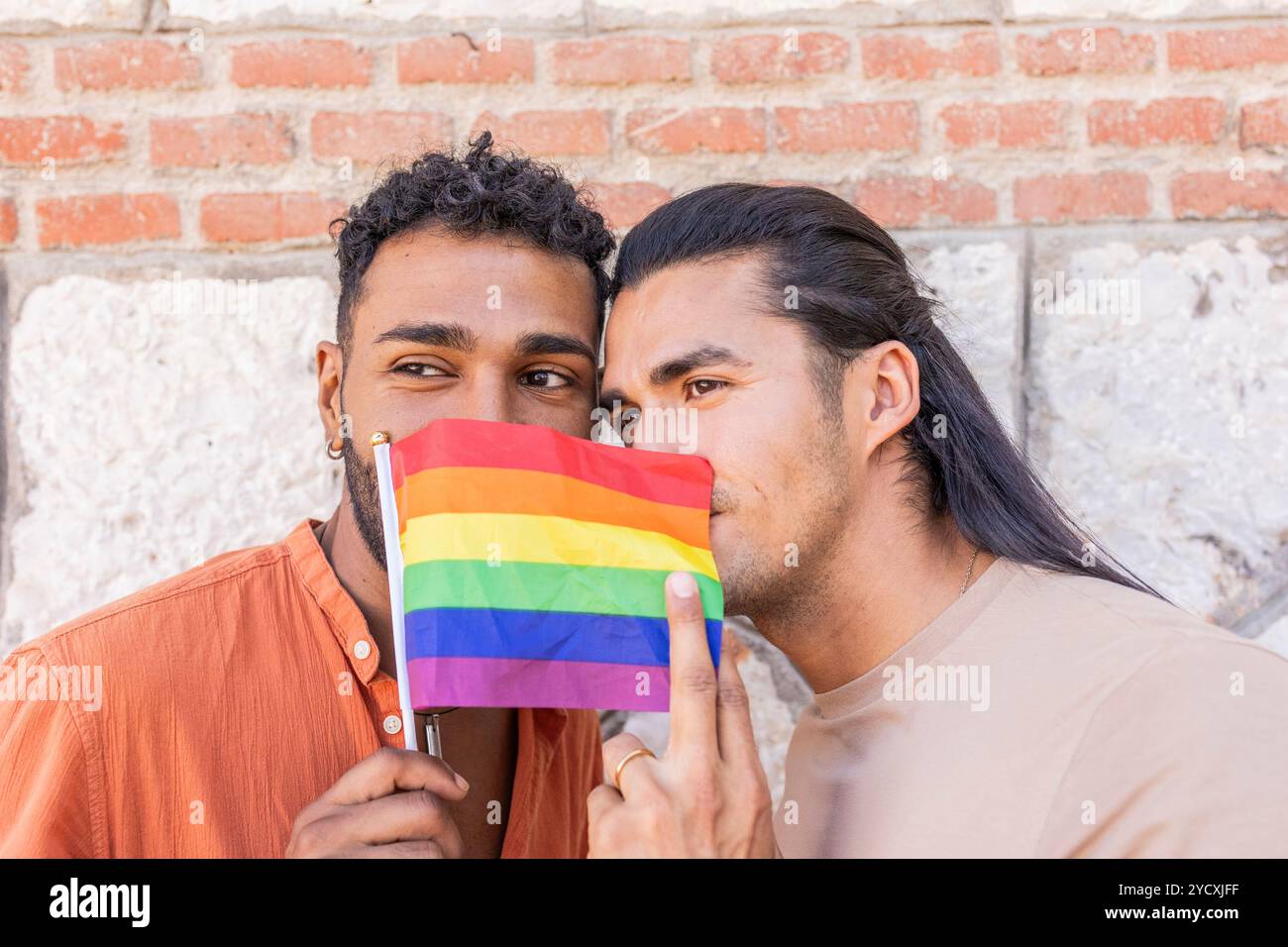 Un couple gay, un Afro-américain et un homme hispanique, partagent un moment tendre tout en embrassant et tenant un drapeau de fierté dans un environnement urbain, Captur Banque D'Images