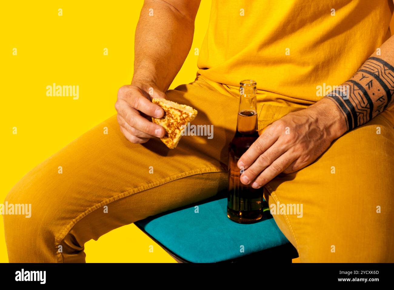 Une scène vibrante mettant en scène un homme vêtu d'un costume jaune, tenant un morceau de tortilla espagnole et une bouteille de bière, contre un dos jaune monochrome Banque D'Images