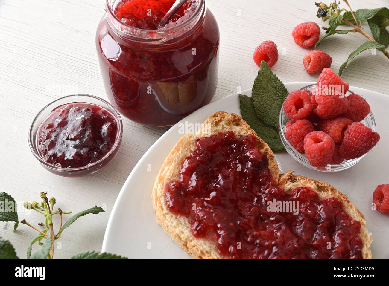 Griller avec de la confiture de framboises sur une assiette blanche avec un bol plein de baies et des pots de confiture sur un banc en bois. Vue surélevée de dessus. Banque D'Images