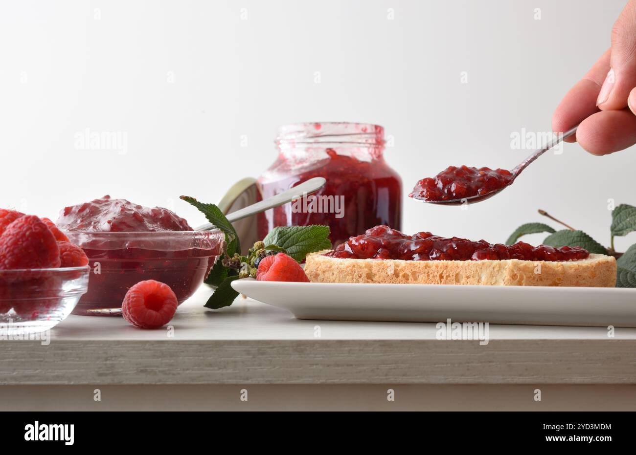 Personne préparant des toasts avec de la confiture de framboises sur une assiette blanche pour le petit déjeuner à la table de la cuisine avec des pots pleins de confiture et un bol avec des baies. Vue de face. Banque D'Images