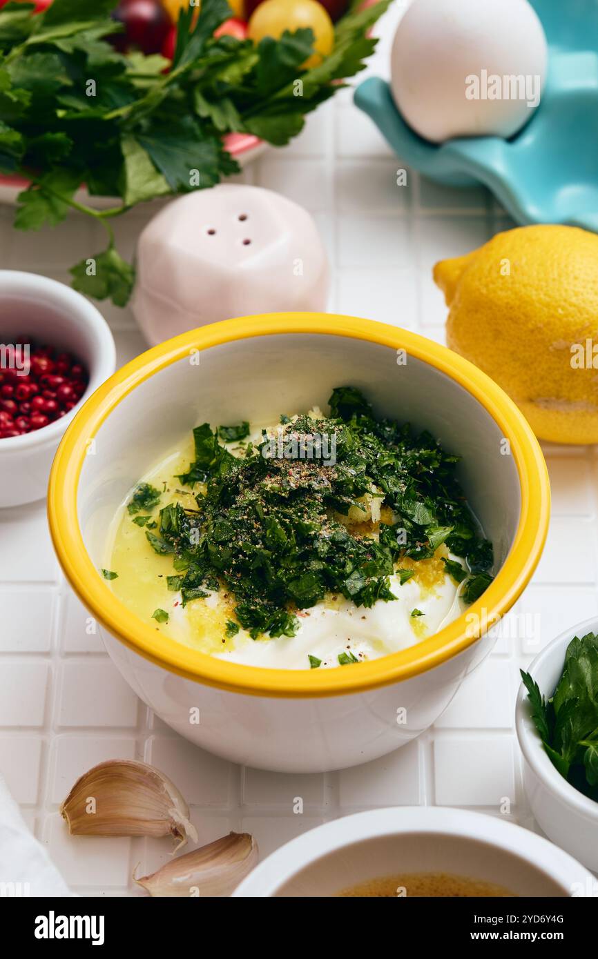 Sauce au yaourt grec avec citron, ail et persil dans un bol blanc avec un bord jaune sur une table blanche avec des ingrédients disposés a Banque D'Images