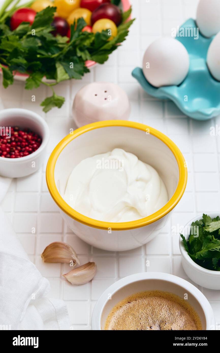 Yaourt grec dans un bol blanc avec un bord jaune sur une table blanche avec des ingrédients disposés autour, salière, citron, herbes, gar Banque D'Images