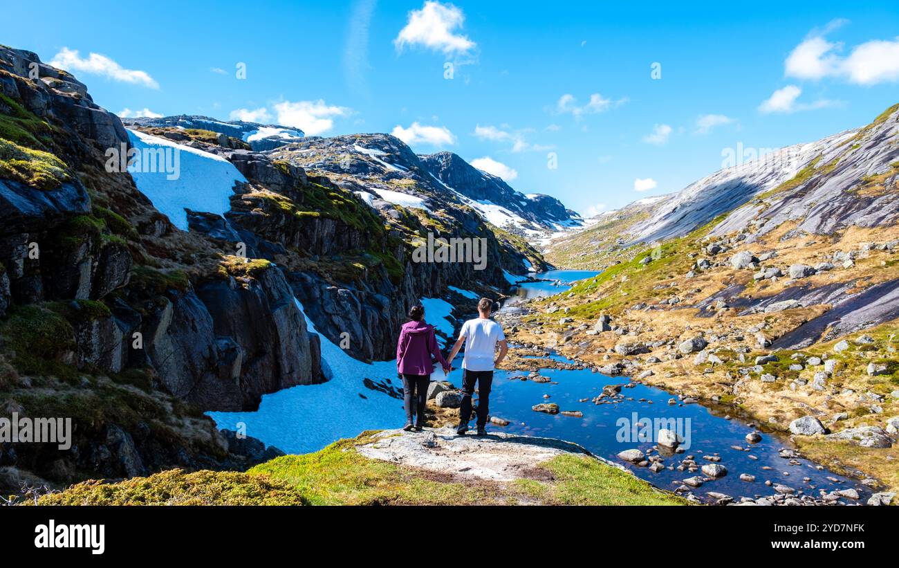 Deux individus se tiennent main dans la main, admirant les paysages époustouflants d'un fjord norvégien, tandis qu'un ruisseau doux serpente à travers les monts accidentés Banque D'Images