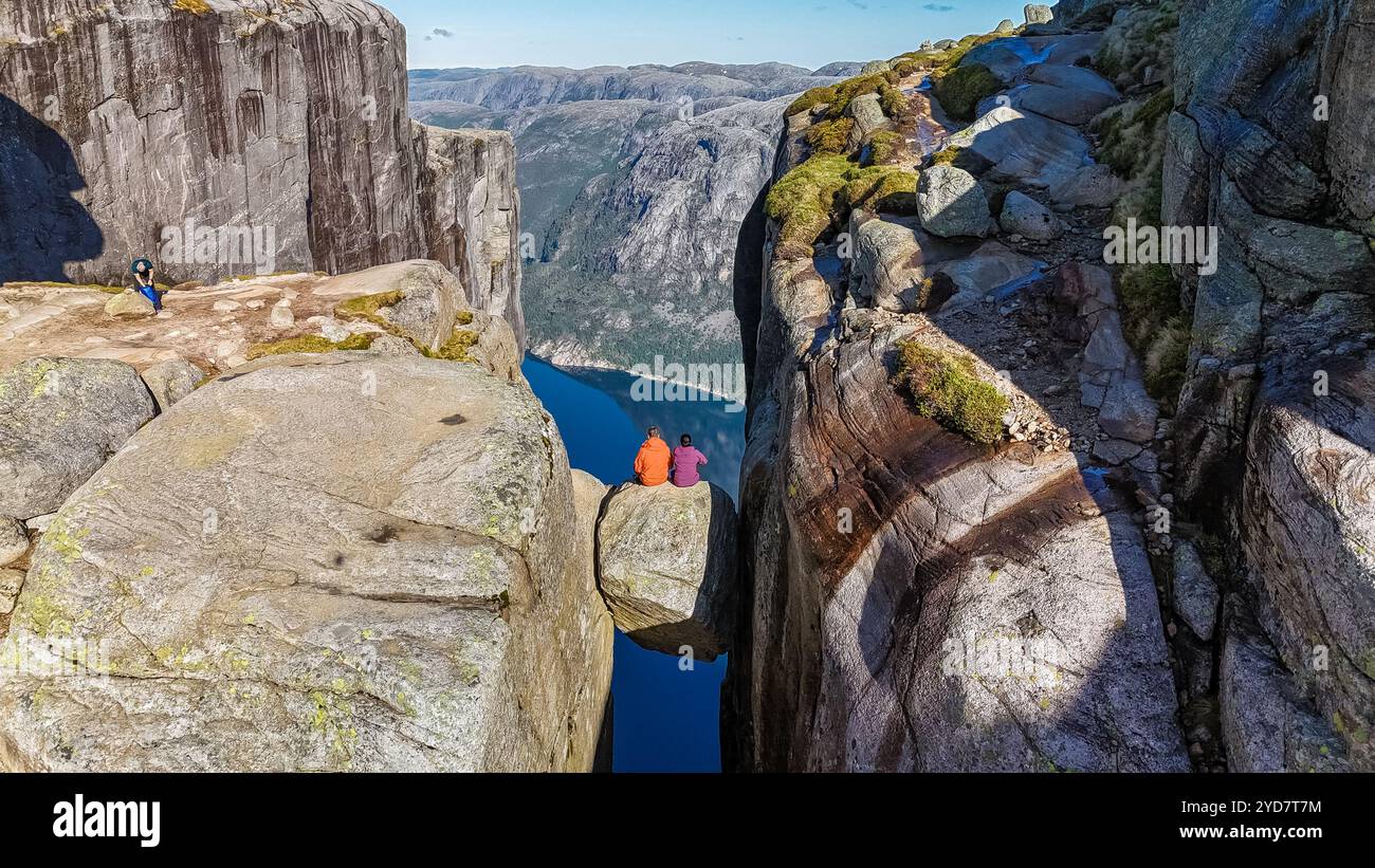 Deux randonneurs sont assis sur le bord de Kjeragbolten Pulpit Rock en Norvège, profitant de la vue panoramique imprenable sur les montagnes environnantes Banque D'Images