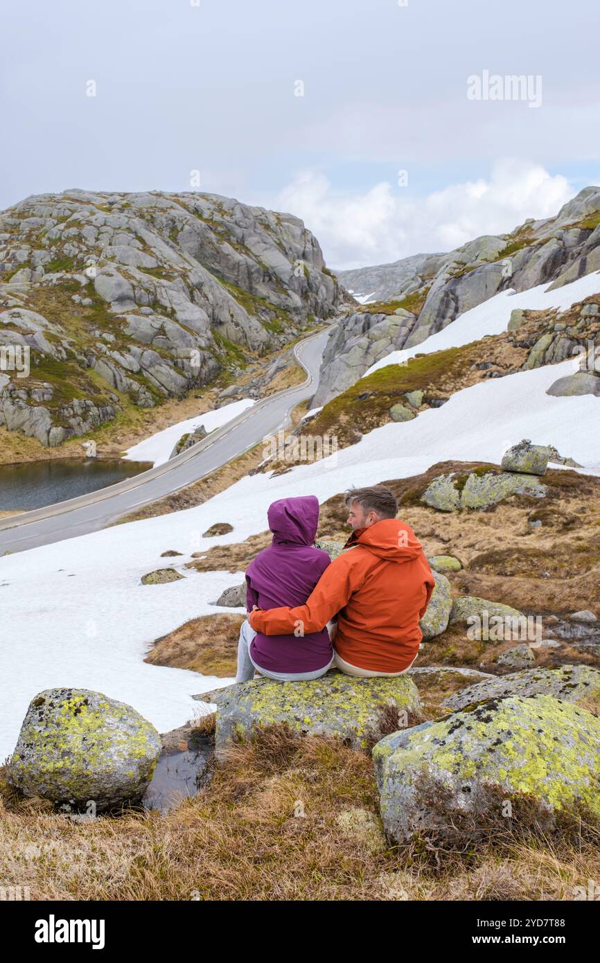 Un couple est assis sur un rocher, profitant de la vue imprenable sur une route sinueuse à travers les montagnes enneigées en Norvège Banque D'Images