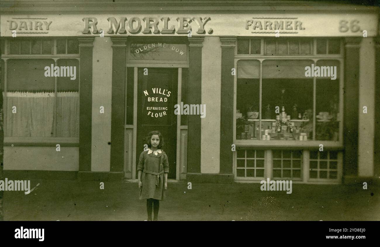 Original WW1 ère carte postale de petite fille debout à l'extérieur dans une porte de magasin. R Morley, Dairy Farmer est le signe du magasin. Les locaux de la laiterie étaient situés au 86, chemin Richmond, Kingston, Surrey. Le panneau de vitrine annonce le pain de Nevill. Circa 1913, Royaume-Uni Banque D'Images