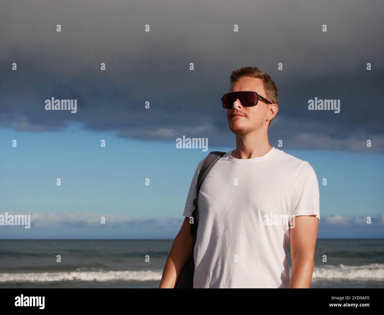 Un jeune homme avec un sac à dos, portant un T-shirt blanc et des lunettes de soleil debout sur le bord de la mer à l'arrière-plan d'un ciel nuageux sombre, avec un espace de copie. Banque D'Images