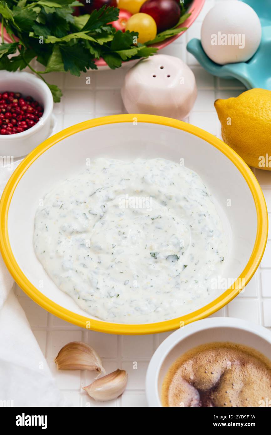 Sauce au yaourt grec avec citron, ail et persil dans un bol blanc avec un bord jaune sur une table blanche avec des ingrédients disposés a Banque D'Images