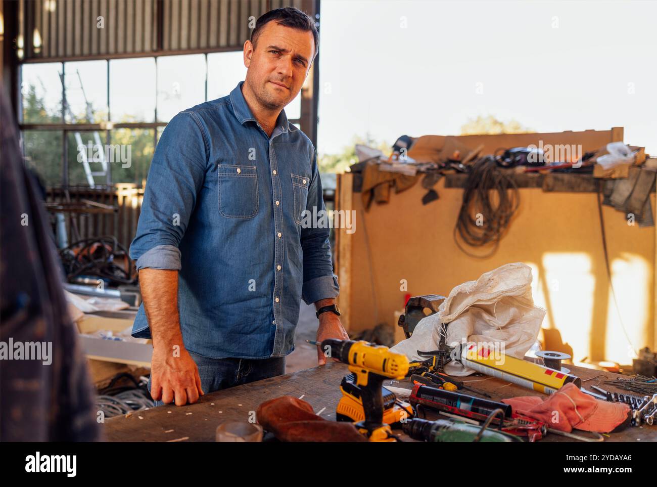 Un jeune homme souriant en vêtements décontractés se tient à une table avec des outils de construction. Banque D'Images