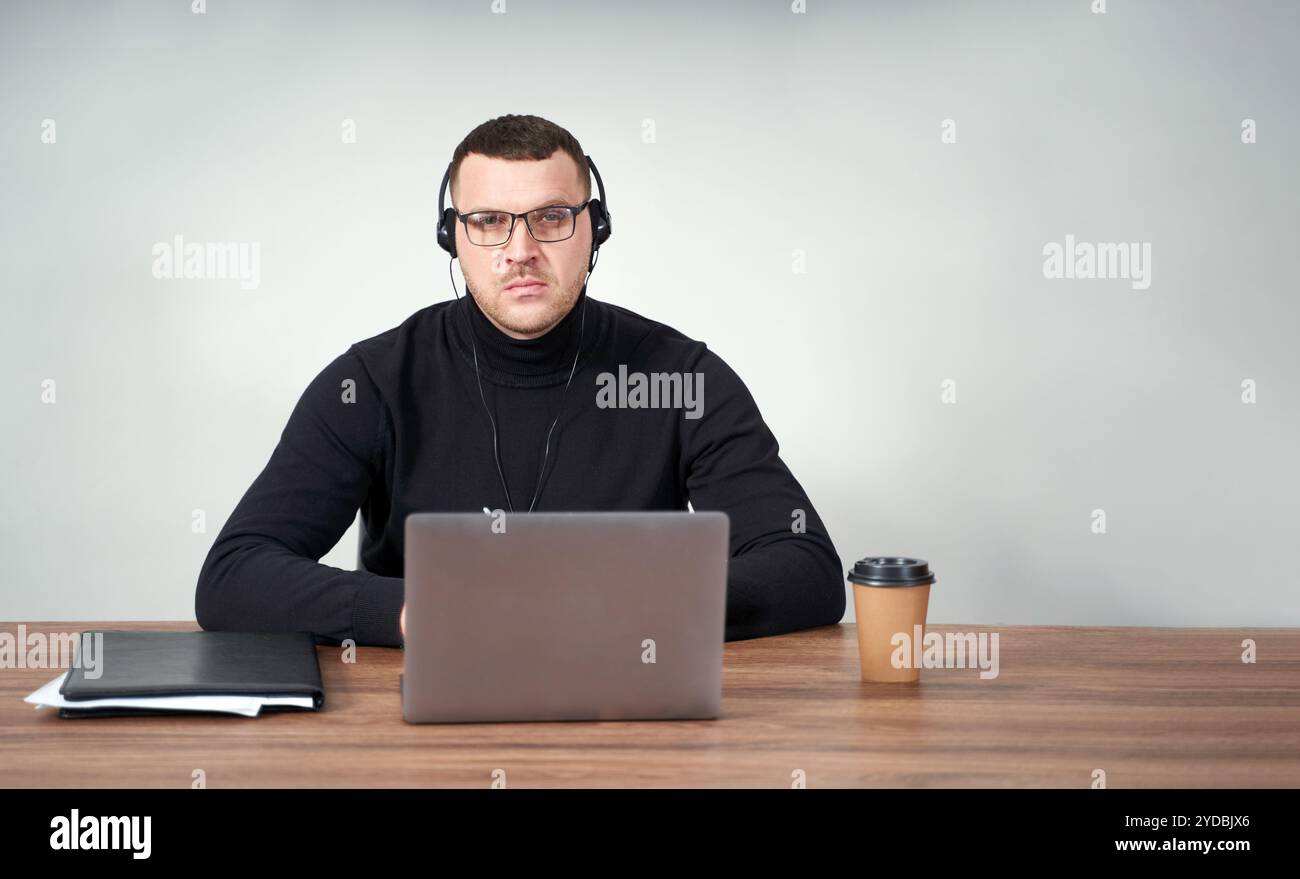 Homme concentré dans des écouteurs à l'aide d'un ordinateur portable tout en étant assis sur fond gris avec fond Banque D'Images