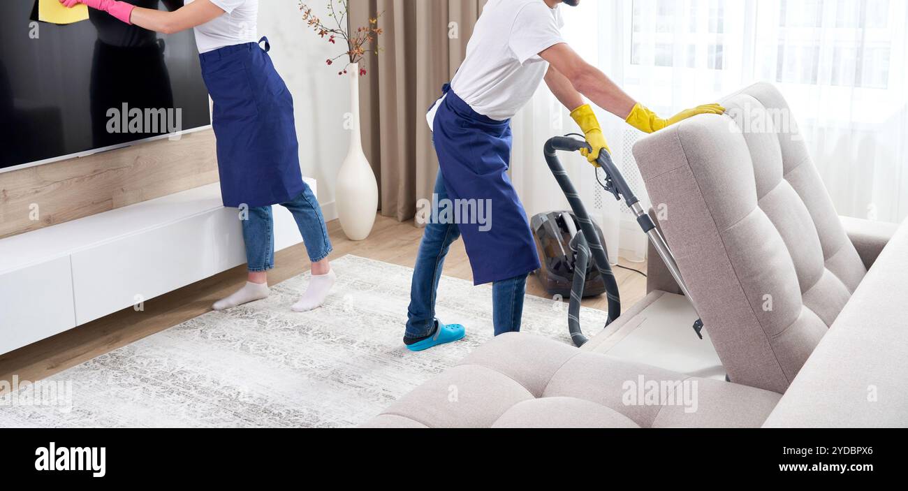 Nettoyants professionnels en sol bleu uniforme et essuyant la poussière des meubles dans le salon de l'appartement. Clea Banque D'Images