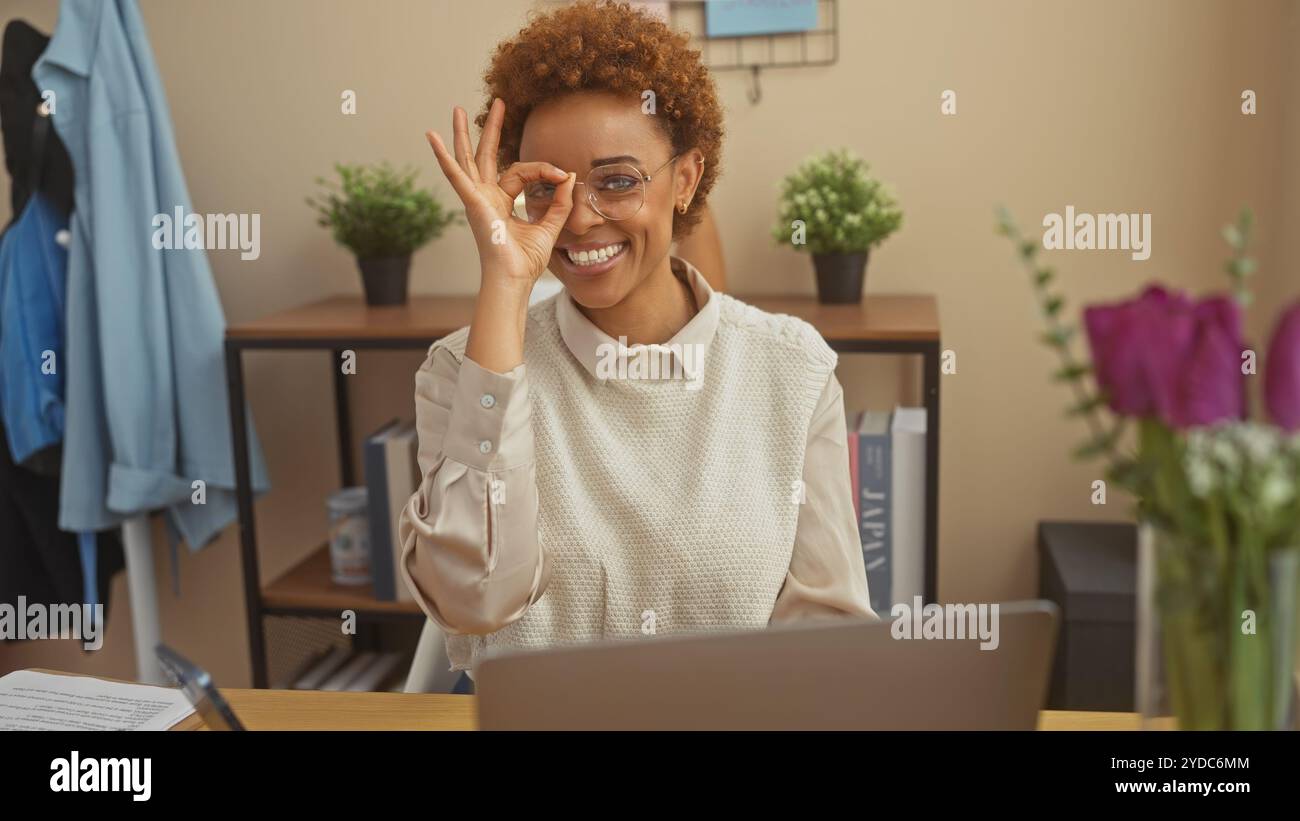 Femme afro-américaine souriante gestuelle signe 'OK' au bureau à domicile avec ordinateur portable, style décontracté, lunettes et plantes. Banque D'Images