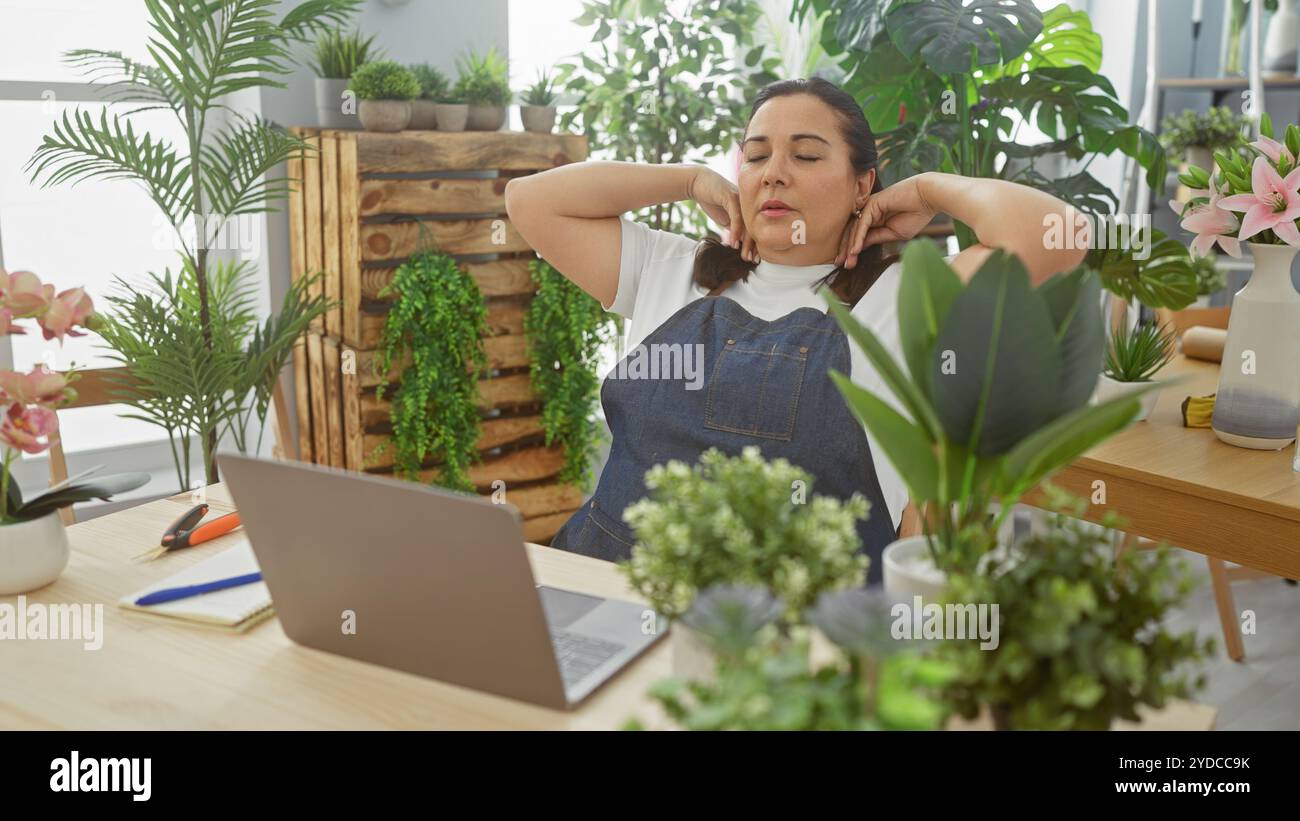 Une femme hispanique d'âge moyen se détend dans un fleuriste intérieur luxuriant rempli de verdure et de plantes en pot, mettant en valeur un environnement d'affaires serein. Banque D'Images