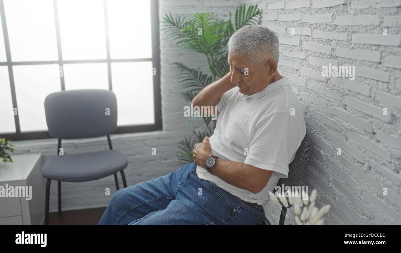 Un homme hispanique aux cheveux gris en chemise blanche est assis dans une salle d'attente intérieure lumineuse, tenant son cou dans l'inconfort à côté d'une chaise moderne et d'un pl vert Banque D'Images