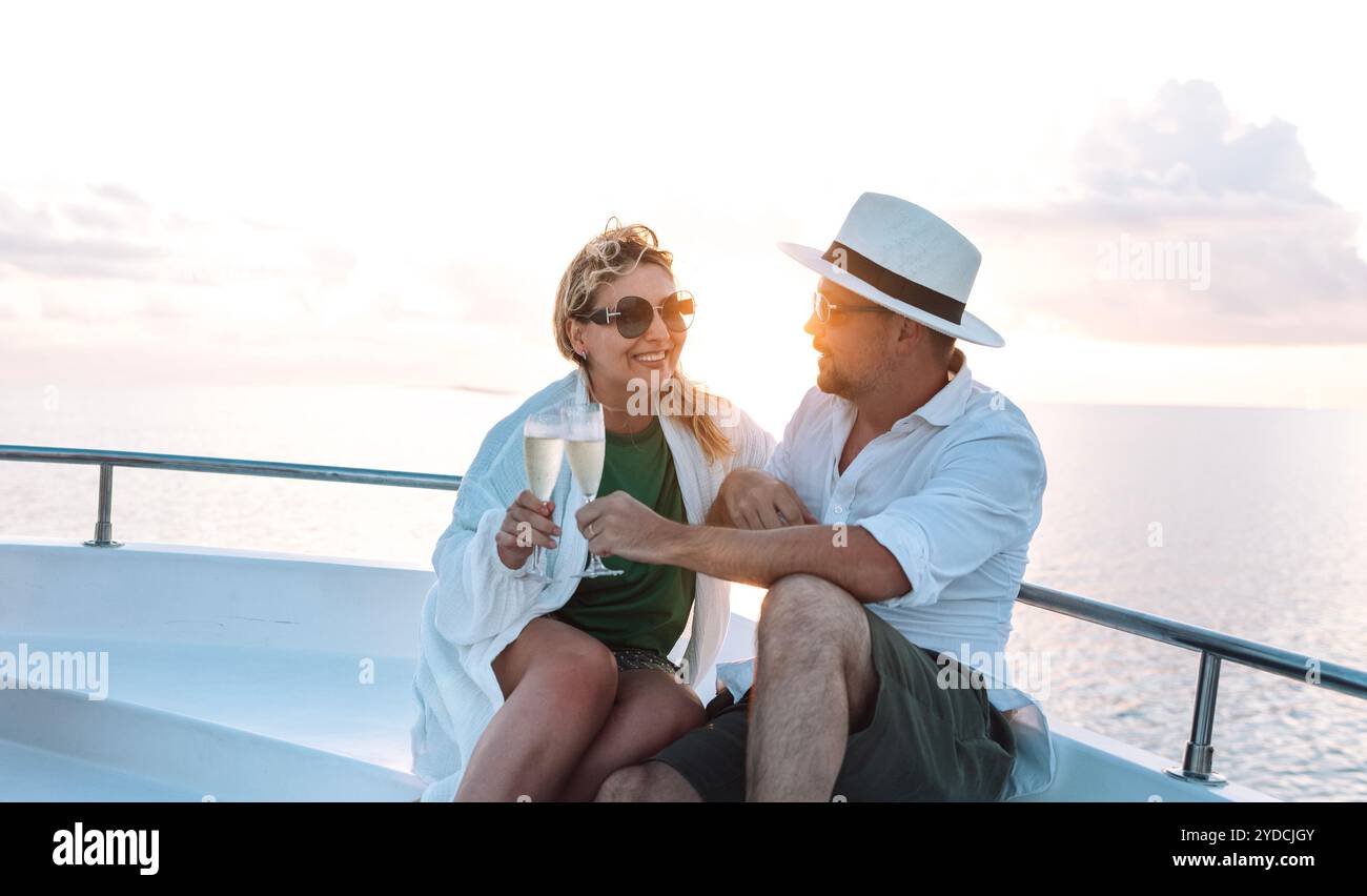 Un jeune couple marié rencontre le coucher du soleil à la poupe du navire. Une femme souriante en lunettes de soleil prit des verres de champagne avec son mari. Un homme an Banque D'Images