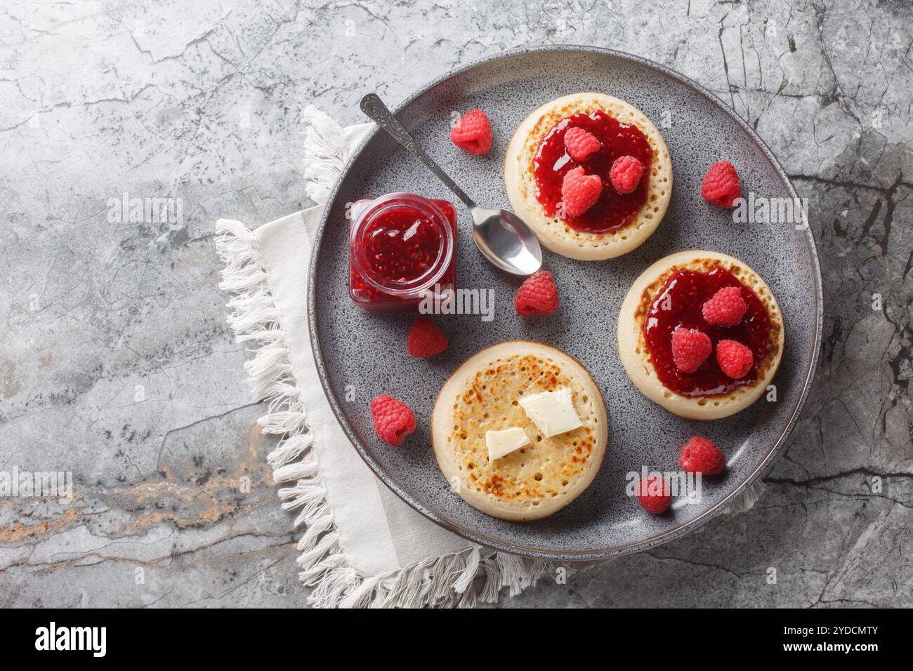 Petit déjeuner de crumpets britanniques avec beurre et confiture de framboises en gros plan sur l'assiette sur la table. Vue horizontale de dessus Banque D'Images