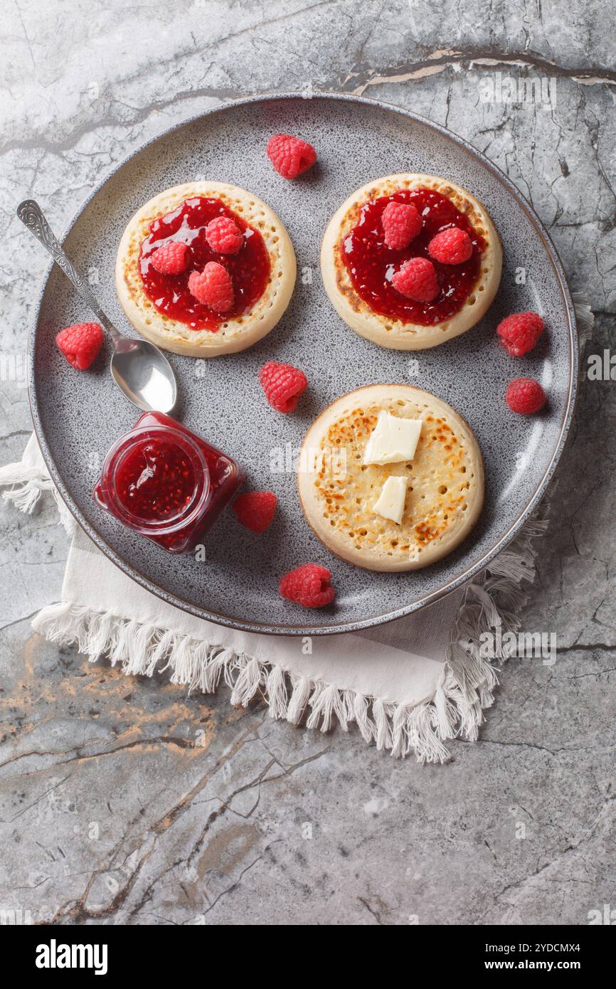 Crumpets grillés chauds avec morceau de beurre et confiture de framboises gros plan sur l'assiette sur la table. Vue de dessus verticale Banque D'Images