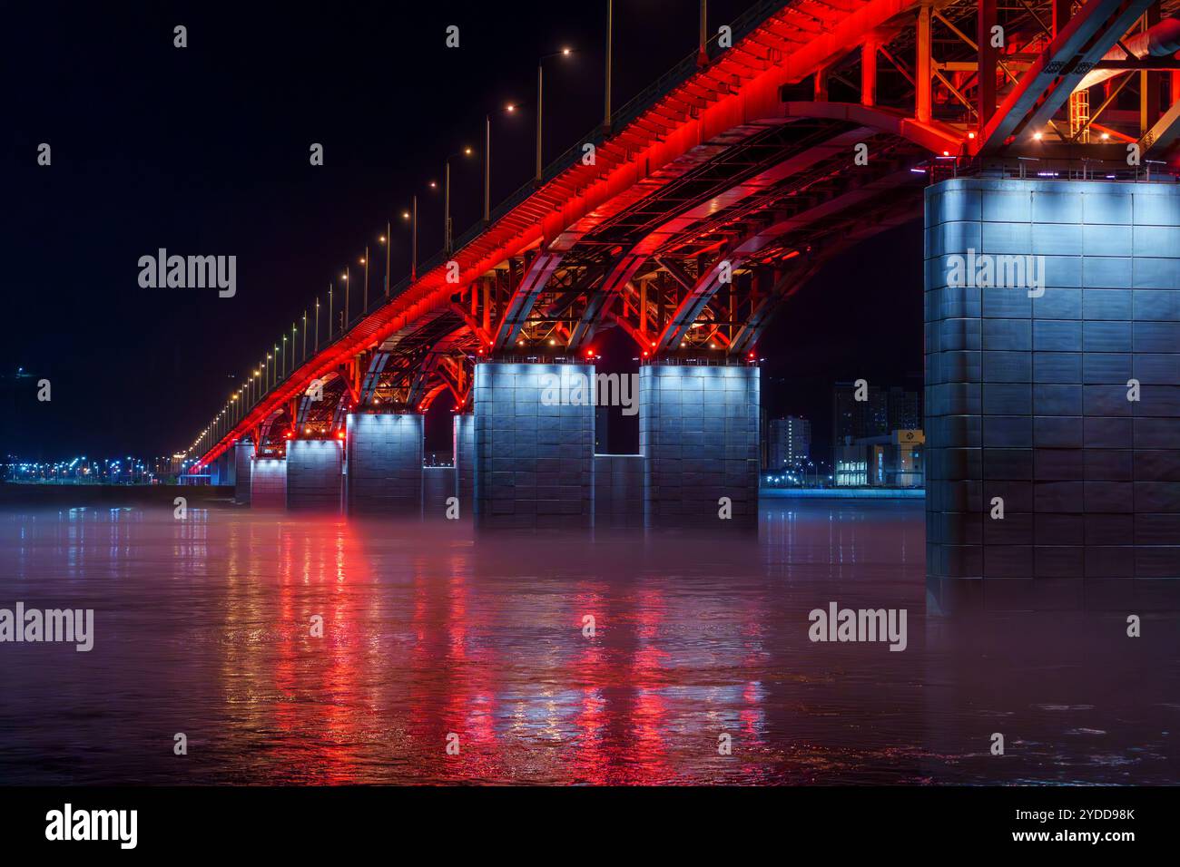 Un pont illuminé rouge s'étend sur une rivière calme la nuit, avec de la brume flottant au-dessus de l'eau et des reflets colorés. La casquette de scène urbaine sereine Banque D'Images