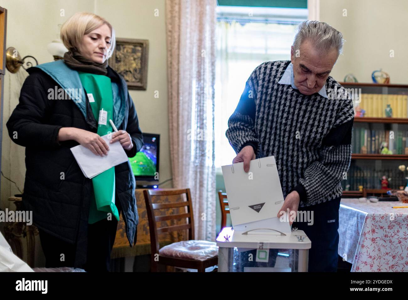 Tbilissi, Géorgie. 26 octobre 2024. Yuri place soigneusement son bulletin de vote rempli dans l'urne mobile du bureau de vote, tenue par un fonctionnaire électoral. Nani observe tranquillement, ses mains reposant sur sa béquille, alors qu'ils achèvent cet important rituel de vote qui représente leur espoir pour l'avenir pacifique et prospère de la Géorgie. (VX photo/ Vudi Xhymshiti) crédit : VX Pictures/Alamy Live News Banque D'Images