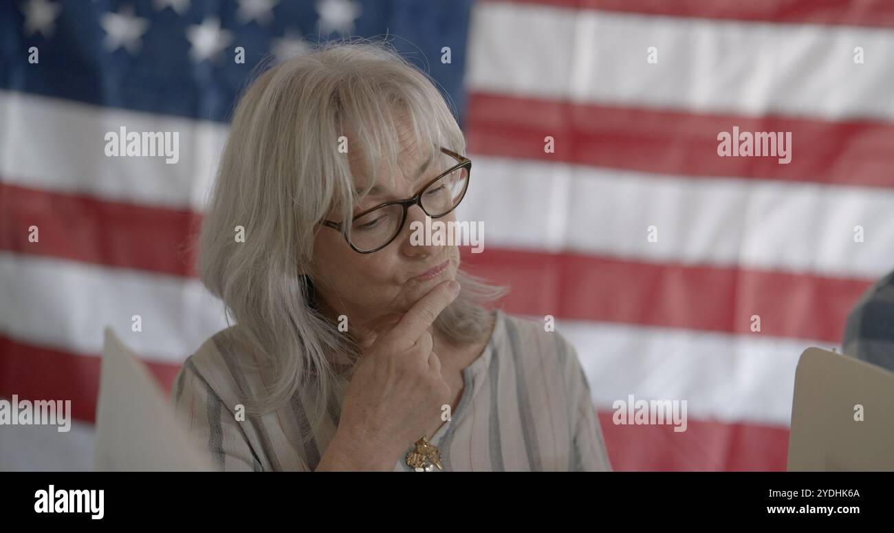 Gros plan sur la femme senior, lunettes et cheveux vers le bas, examine les options tout en votant au stand, avec le drapeau des États-Unis sur le mur derrière Banque D'Images Gros plan sur la femme senior, lunettes et cheveux vers le bas, examine les options tout en votant au stand, avec le drapeau des États-Unis sur le mur derrière Banque D'Images