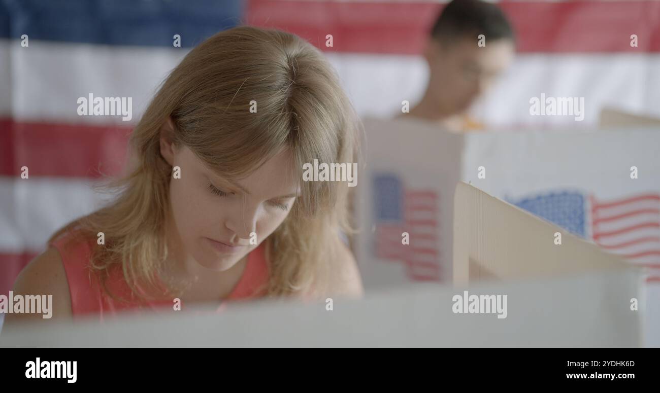 Gros plan moyen sur une jeune femme aux cheveux roux regarde vers le bas tout en votant au stand et en considérant les candidats. L'homme asiatique vote derrière elle Banque D'Images Gros plan moyen sur une jeune femme aux cheveux roux regarde vers le bas tout en votant au stand et en considérant les candidats. L'homme asiatique vote derrière elle Banque D'Images