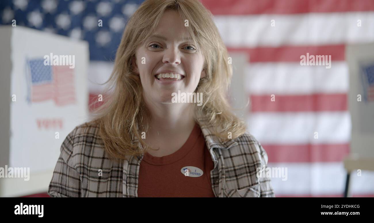 Une jeune femme aux cheveux roux porte l'autocollant "J'ai voté" et sourit. Cabines de vote visibles derrière elle, et drapeau des États-Unis sur le mur arrière en arrière-plan Banque D'Images Une jeune femme aux cheveux roux porte l'autocollant "J'ai voté" et sourit. Cabines de vote visibles derrière elle, et drapeau des États-Unis sur le mur arrière en arrière-plan Banque D'Images