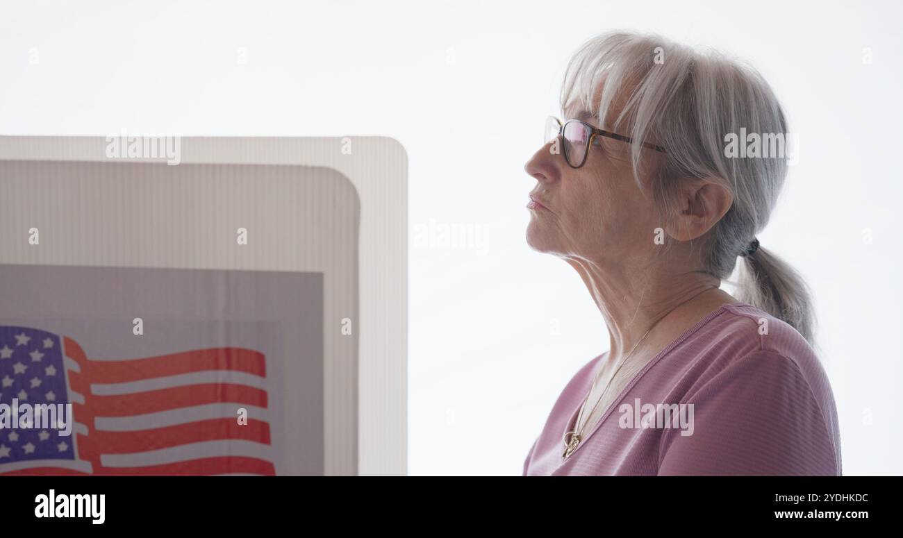 Femme senior avec des lunettes complète le bulletin de vote à la cabine de vote, avec fond blanc derrière. Gros plan Banque D'Images Femme senior avec des lunettes complète le bulletin de vote à la cabine de vote, avec fond blanc derrière. Gros plan Banque D'Images