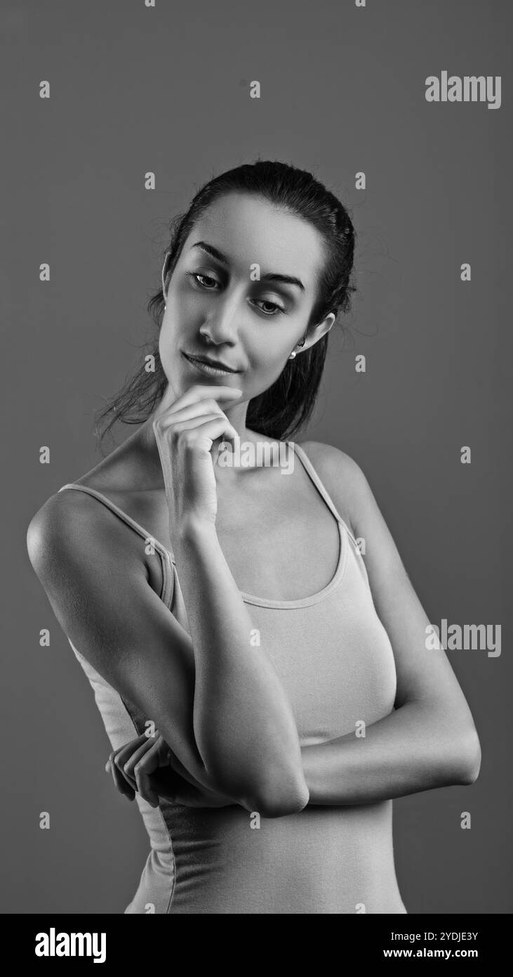 Jeune femme en débardeur, main sur le menton, regardant réfléchi en noir et blanc portrait sur fond gris Banque D'Images