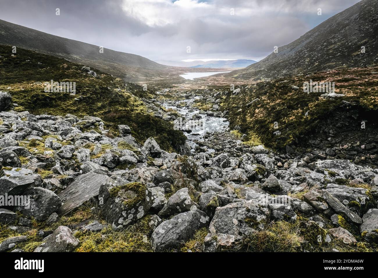 Une vallée de montagne rocheuse avec un petit ruisseau qui la traverse vers un lac au loin. Le ciel est couvert. Les pentes sont couvertes d'herbe Banque D'Images