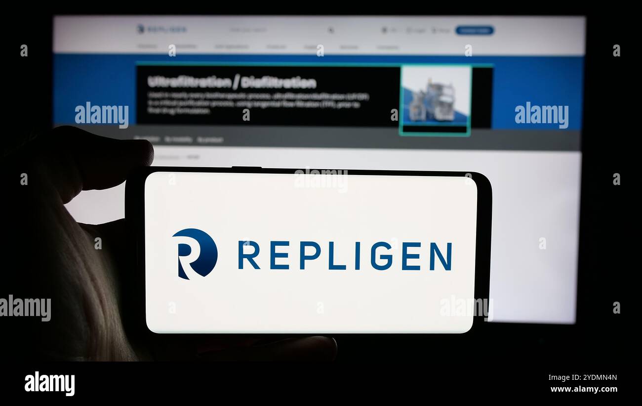 Personne tenant le téléphone portable avec le logo de la société pharmaceutique américaine Repligen Corporation sur l'écran en face de la page Web. Concentrez-vous sur l'affichage du téléphone. Banque D'Images