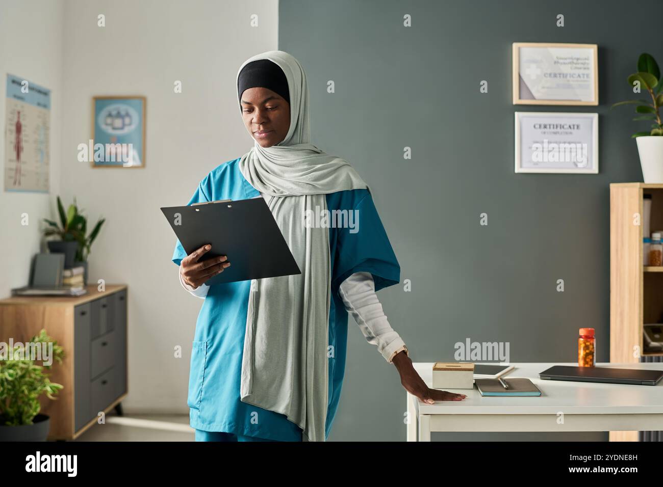 Professionnel de la santé musulman portant des gommages et debout dans un cadre de bureau moderne, examinant des cartes, avec des certificats et des diplômes sur le mur et divers articles de bureau autour Banque D'Images
